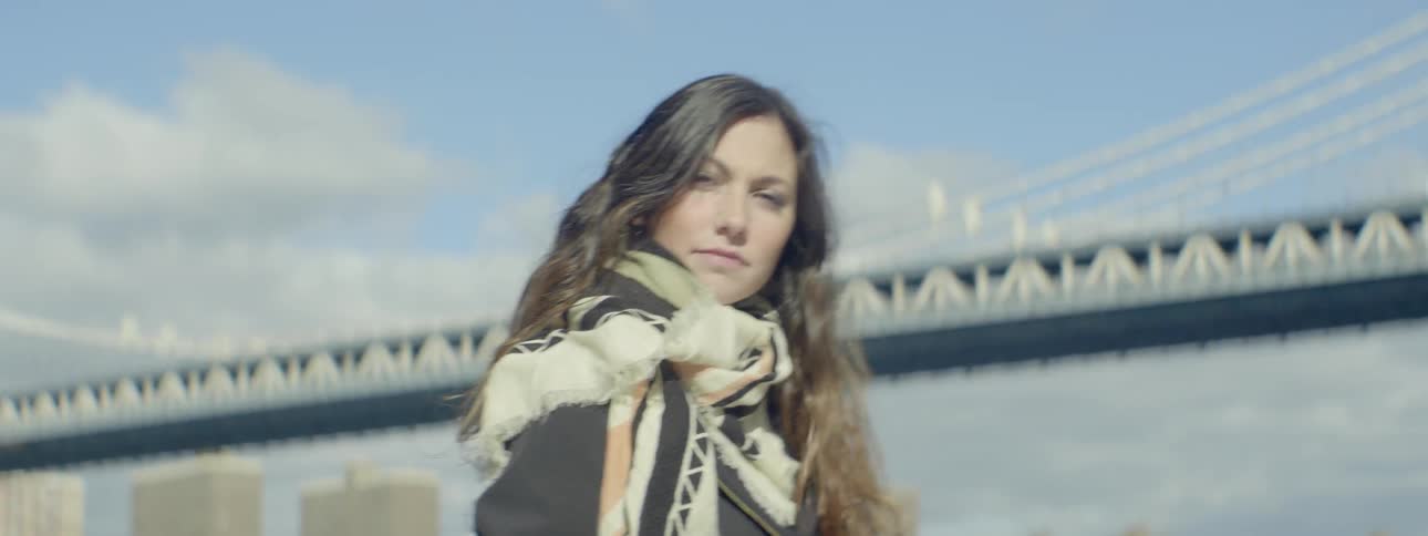 A woman stands in front of a bridge with a city in the background