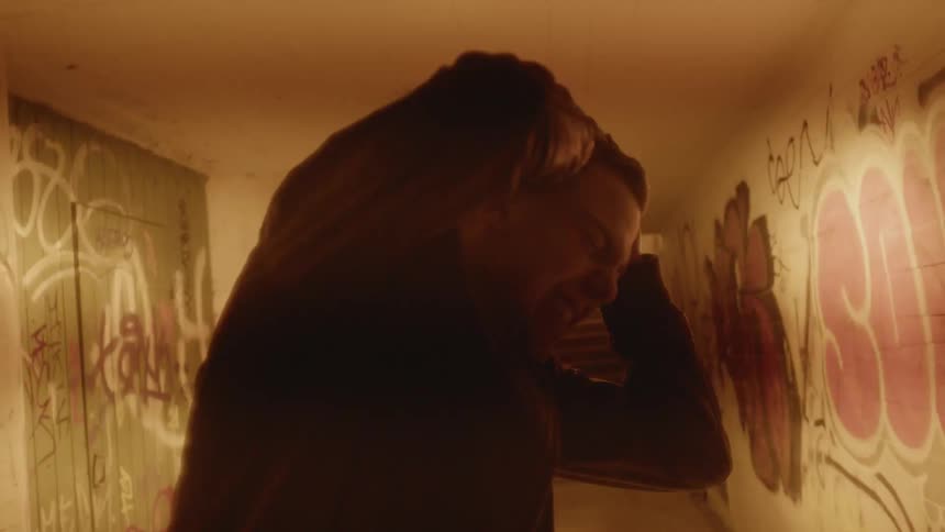 Young man holds his head in pain while standing inside a dimly lit hallway