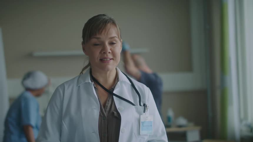 Doctor steps forward with smile to give next of kin happy news and nurses assist patient lying in hospital bed in background