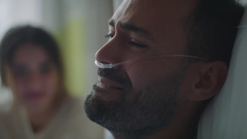 A man laying in a hospital bed in a hospital room next to a woman with a wall and window in the background