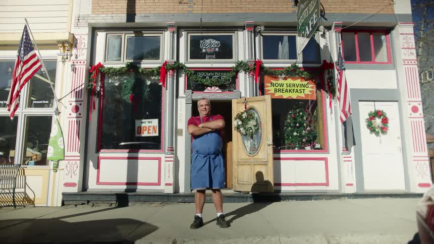Man wearing red shirt and blue apron stands in front of cafe with christmas decorations crossing his arms