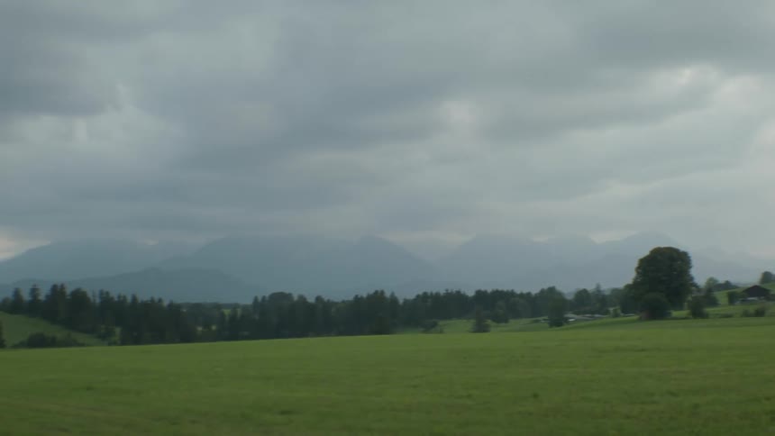 Fog obscures mountains in background as conifer trees dot hills and area near buildings bordering grassy fields