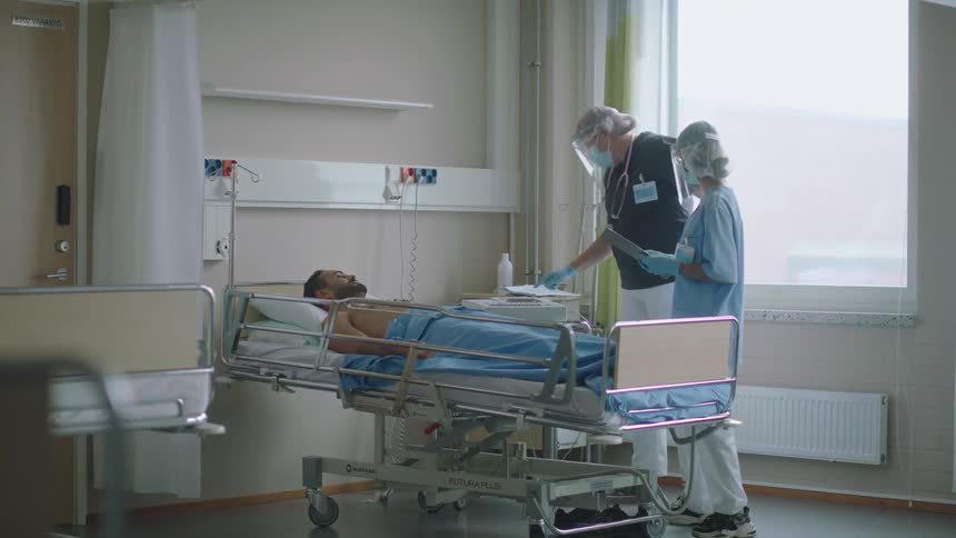 Medical personnel consult patients chart as male patient lies in hospital bed watching them