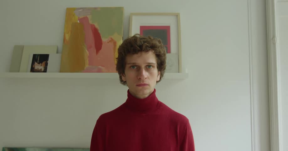 Man with curly hair and red turtle neck sweater blinks at camera