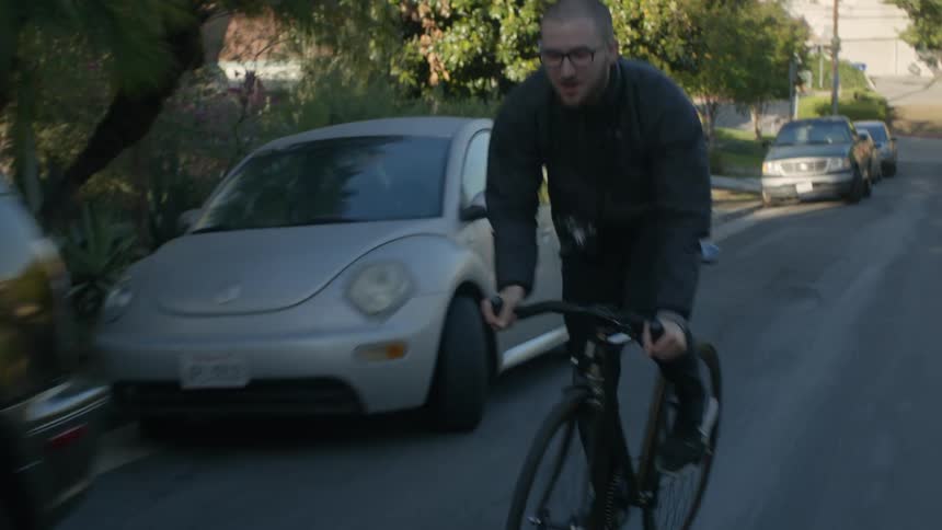 A bald man with glasses and a camera bikes up a hill on a residential street with some effort