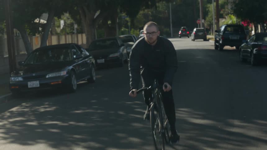 A man is riding a bicycle along a neighborhood street lined with parked vehicles in the daytime