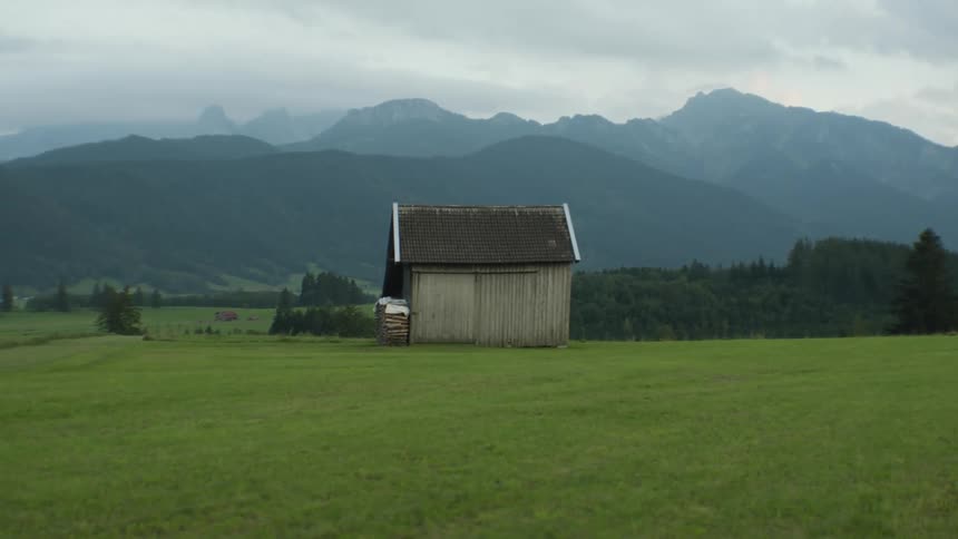 Building stands on field near forest with fog covered mountains in background under a cloudy sky