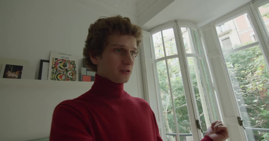 A young man stands in a house and smiles and talks with someone as he makes actions with his arms and hands like he is playing a musical instrument