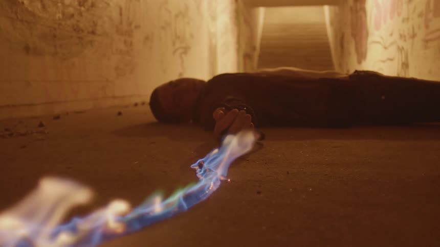 A man is lying unconscious on the floor in a house holding an electrical cord in his hand that is on fire