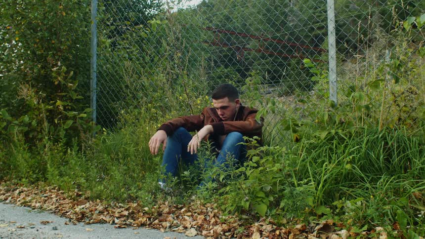 Man wearing jacket sits on ground among vegetation near chain link fence looking down