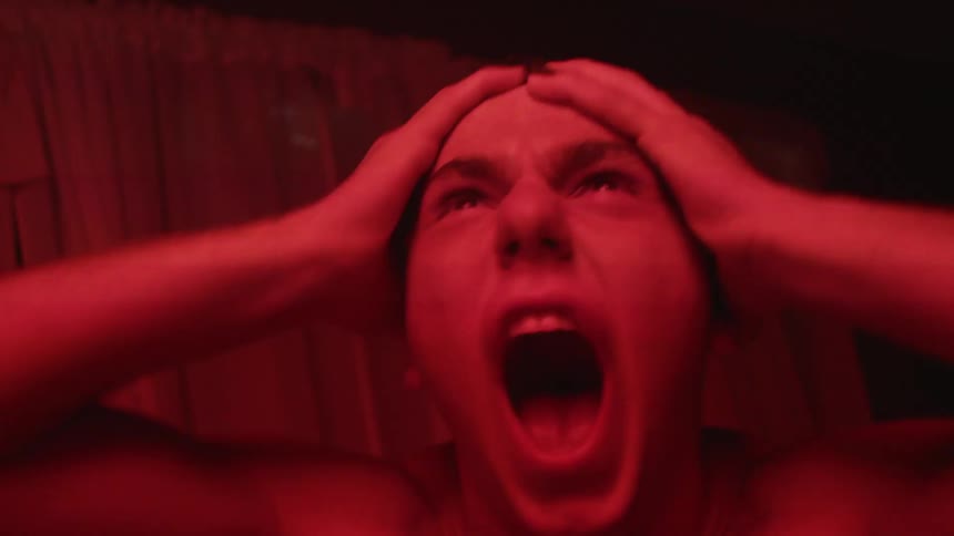 Young shirtless man with hands over the head screaming inside a room under red strobing lights