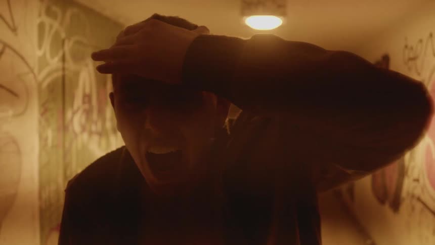 A man holding his head in a hallway with graffiti on the walls with stairs in the background