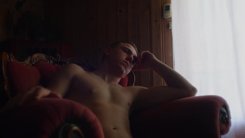 A shirtless man sits in a chair and rests his head on his hand