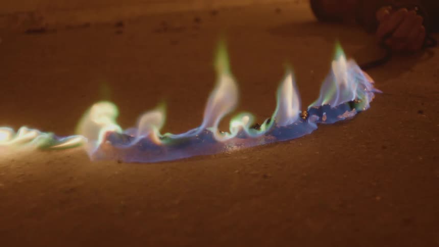 A young man lies in the sand with his hands tied to a rope that is on fire and quickly approaching him