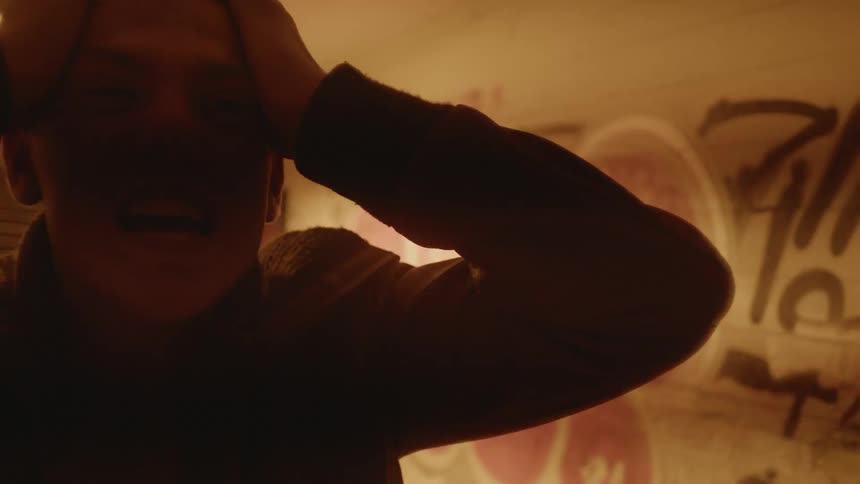 A man holds his head while screaming in a room tagged with graffiti