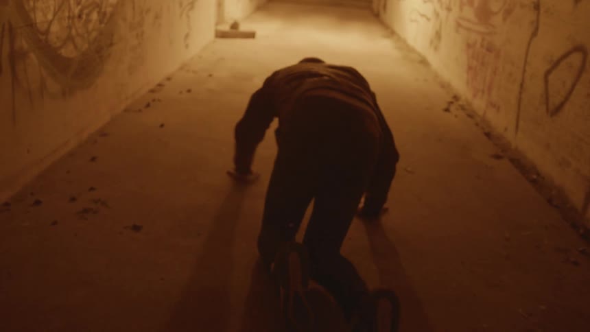 A man stumbles to the ground and crawls along a dim hallway marked with graffiti