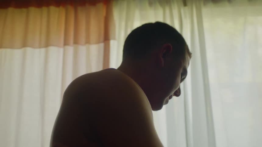 Bare chested man standing near curtained window with arms wrapped around himself jerks to the side