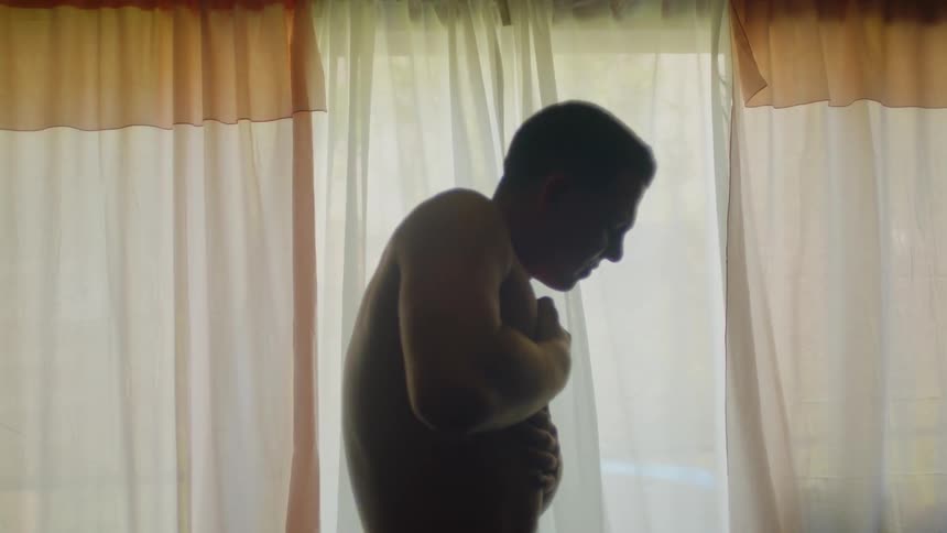 Young muscular shirtless man shakes while rubbing his chest inside a dimly lit room