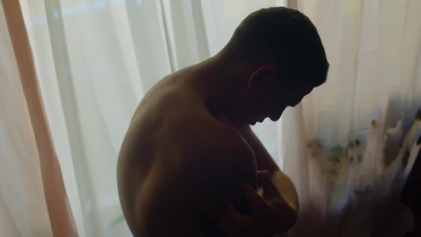 Young shirtless man standing inside a dimly lit room at home rubbing his arms and shoulder