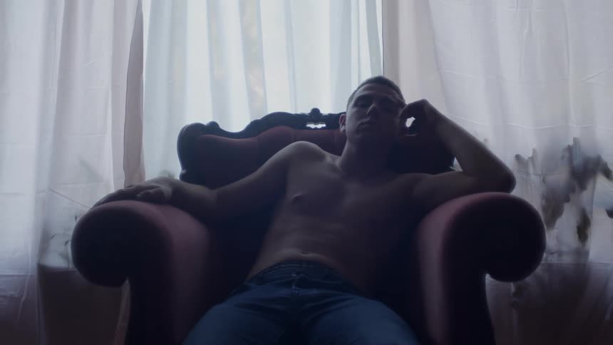 A man in a room sitting on a couch with a window and curtains in the background
