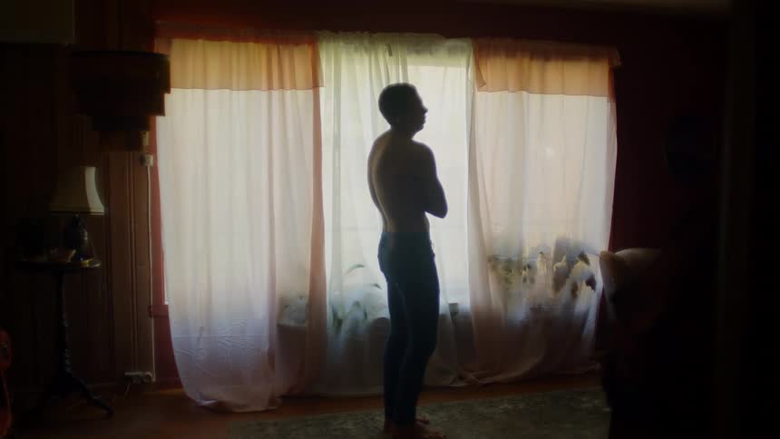 Silhouette of a young shirtless man standing in front of a window at home