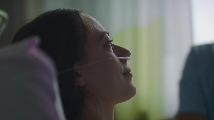 A woman laying in a hospital bed with an oxygen tube in her nose with a person and a window in the background