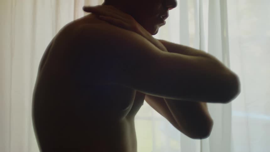 Young shirtless man rubs his chest inside a dimly lit room near a window