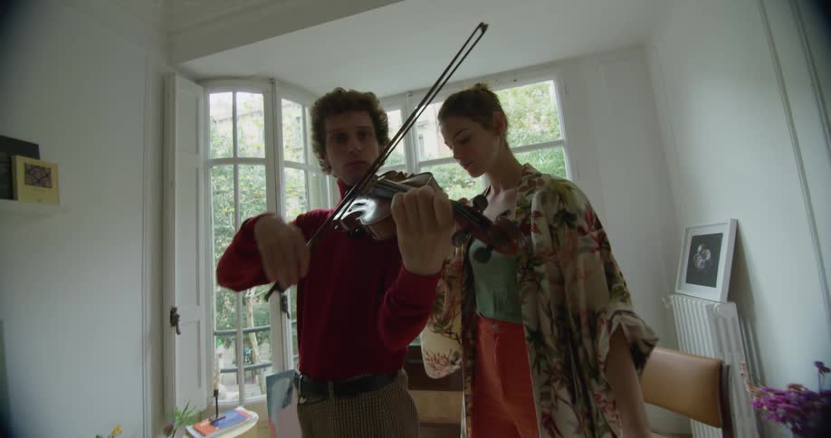 A young woman is standing next to a man who is playing a violin in a living room