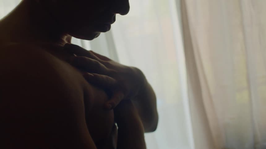 A man runs his hands along his chest as he stands bare chested in front of a window