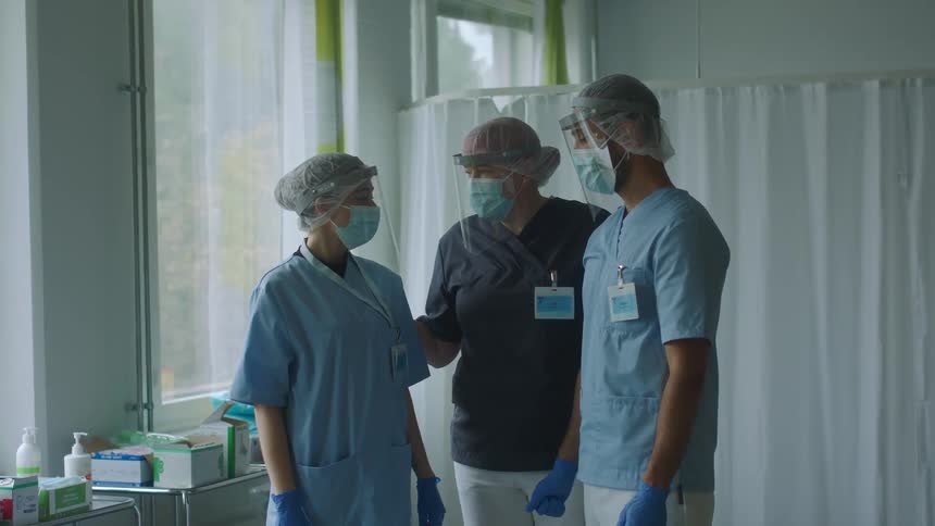 Senior medical nurse comforts tow other medical professional as they stand in protective gear in hospital room near window