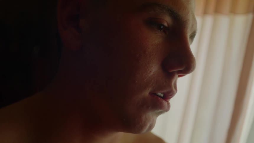 Young shirtless man rubs his own chest standing inside a dimly lit room