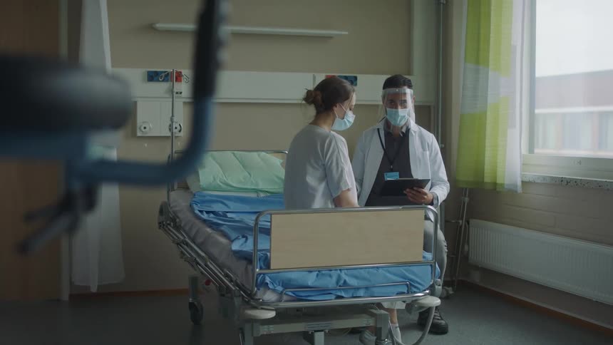 A woman is sitting on a hospital bed as a doctor speaks to her