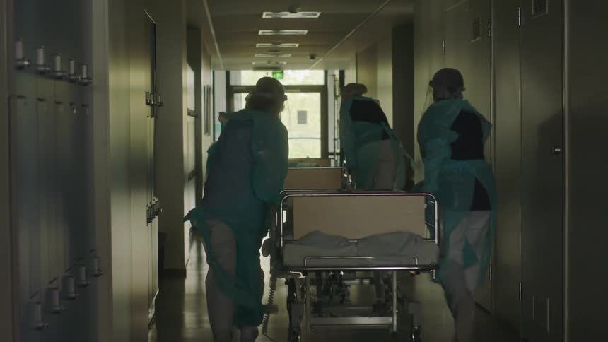 Medical team in protective gear wheel two patients in hospital beds down hallway at hospital