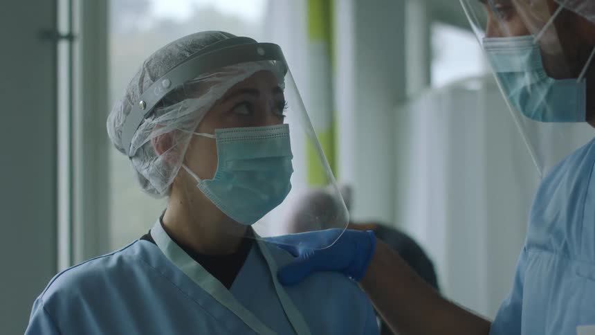 Male nurse comforts female nurse as they stand in hospital room talking