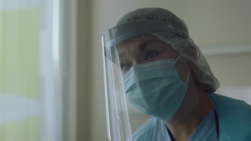 Nurse in scrubs and face shield leans forward to talk before straightening