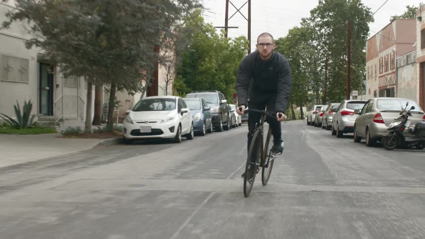 A man pedals a bike down the middle of a neighborhood street with cars parked on both sides