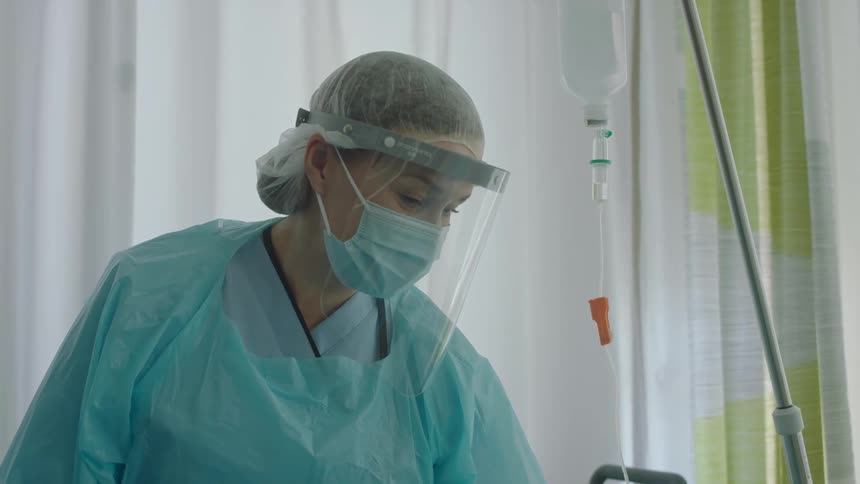 A woman in a hospital room with a mask face shield and scrubs