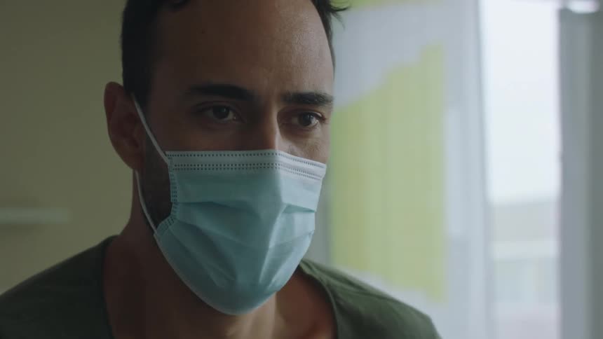 Two nurses with mask and scrubs help in a man to stand in a hospital room with a wall and window in the background
