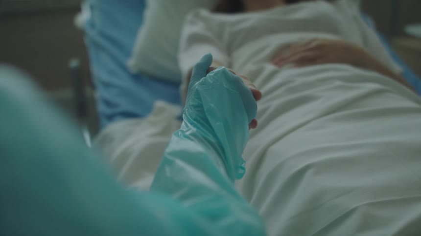 A patient in a hospital bed reaches out to a nurse and the nurse takes her hand to comfort her