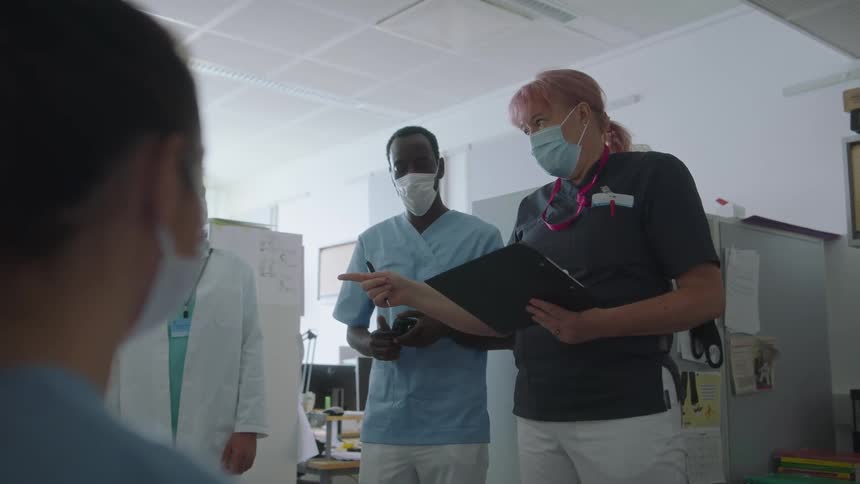 A woman with a mask and a clipboard in a room talking to a doctor and nurses with a wall in the background