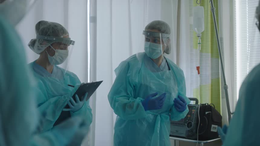 Nurses are dressed in scrubs in a hospital room and one person is writing on a clip board