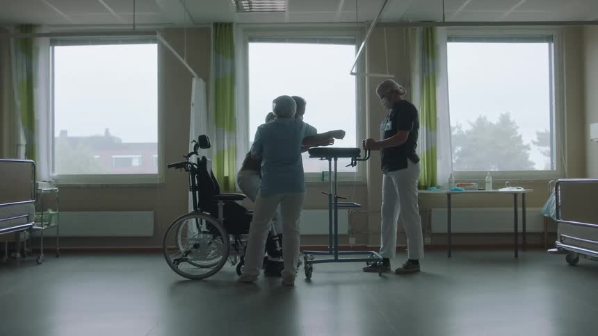 Nurses helping a patient to stand from a wheelchair in a hospital room