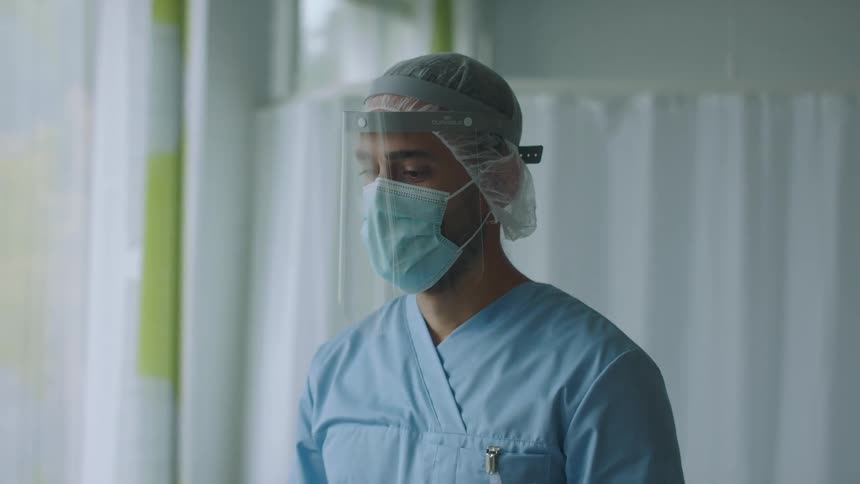 Male nurse wearing scrubs face shield and face mask stands near window in hospital room