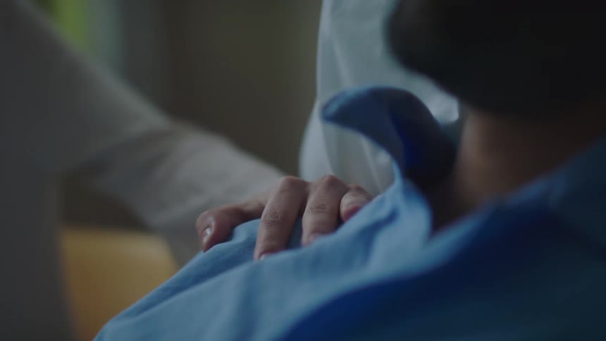 A woman touches the shoulder of a man laying in a hospital bed while fully clothed