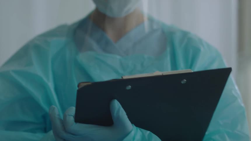 A nurse in personal protective equipment writes on a clipboard while standing in a hospital