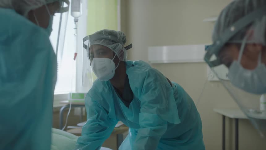 Three nurses with face shields and masks in a hospital room with a wall and window in the background
