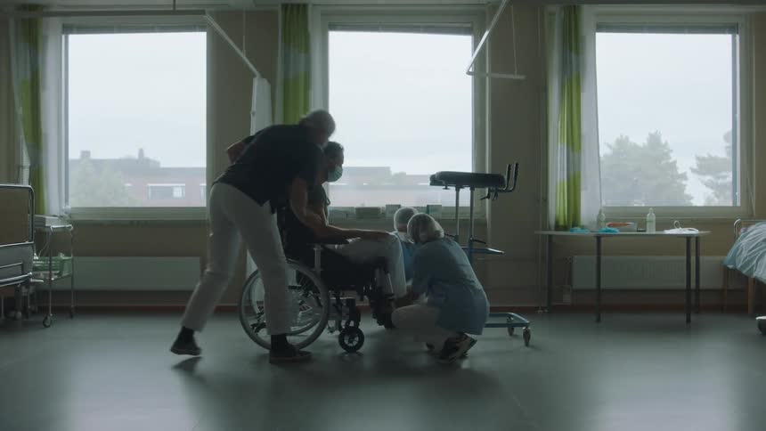 A man in a wheelchair in the hospital room with three nurses with a wall and window in the background