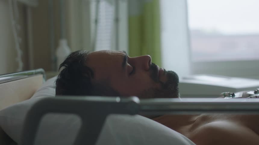 Young bearded man laying still on a hospital bed near a window