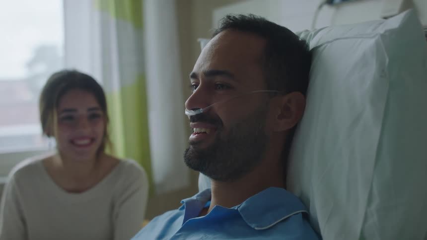 A man laying in a hospital bed in a hospital room next to a woman with a wall and window in the background