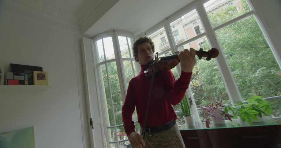 Man wearing long sleeve shirt stands near windows in living room playing violin